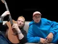 Ed Sheeran z gitarą i młody mężczyzna w niebieskiej bluzie siedzą na kanapie.
