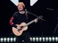 Ed Sheeran gra na gitarze akustycznej na scenie, ubrany w czarną bluzę z czerwonymi pasami na rękawach.