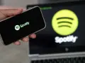 Dłoń trzyma smartfon z logo Spotify. W tle ekran laptopa z tym samym logo.