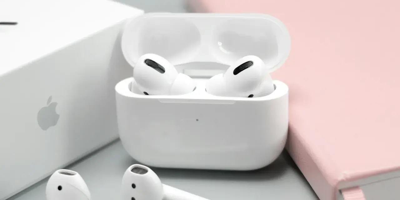 Białe słuchawki AirPods w etui, obok pudełka Apple i różowego notatnika.