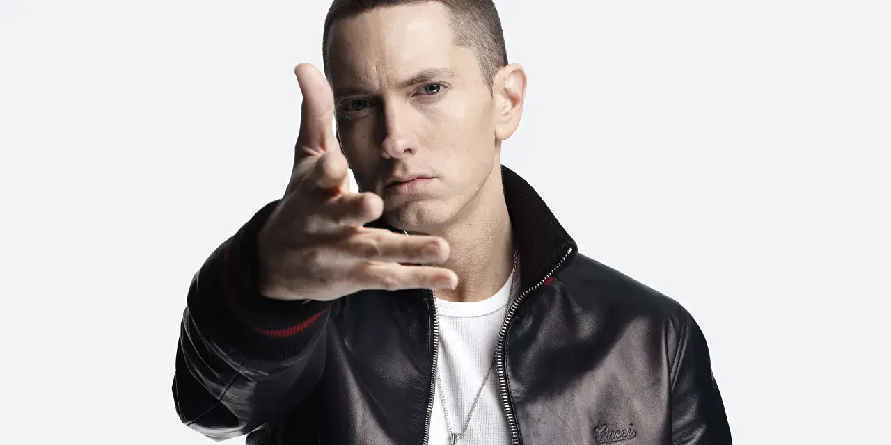 Eminem w czarnej skórzanej kurtce i białej koszulce, z ręką wyciągniętą w stronę widza.