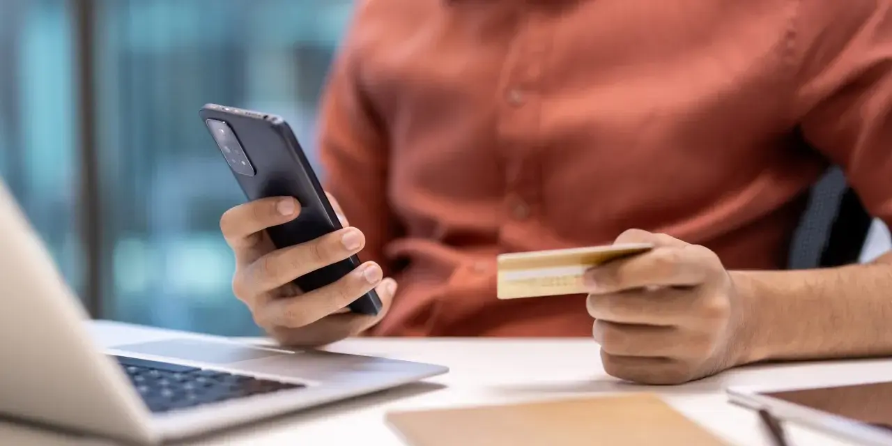 Dłoń trzymająca telefon i kartę kredytową przy laptopie.