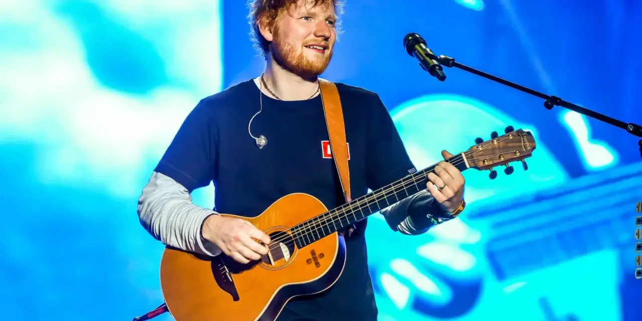 Ed Sheeran gra na gitarze akustycznej na tle niebieskiej sceny.