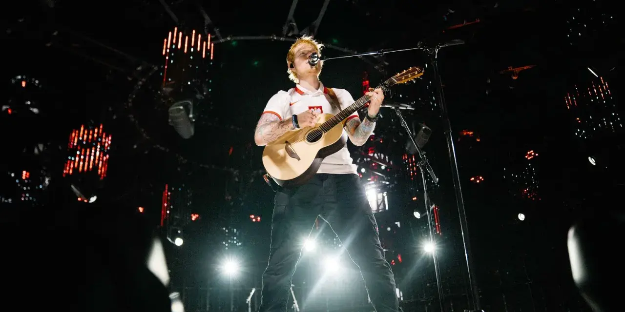 Ed Sheeran gra na gitarze na scenie w białej koszulce z polskim godłem.