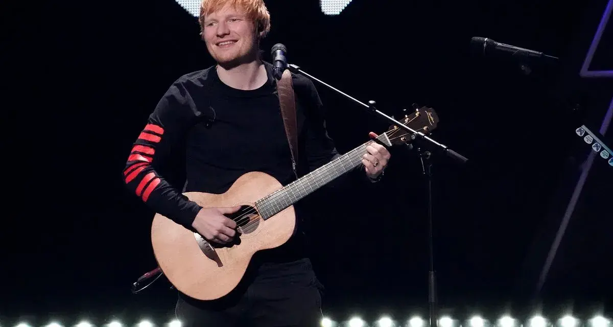 Ed Sheeran gra na gitarze akustycznej na scenie, ubrany w czarną bluzę z czerwonymi pasami na rękawach.