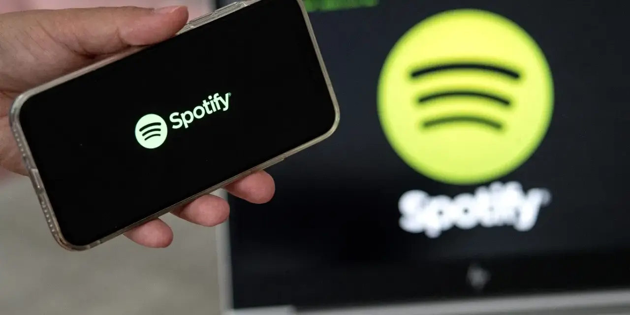 Dłoń trzyma smartfon z logo Spotify. W tle ekran laptopa z tym samym logo.