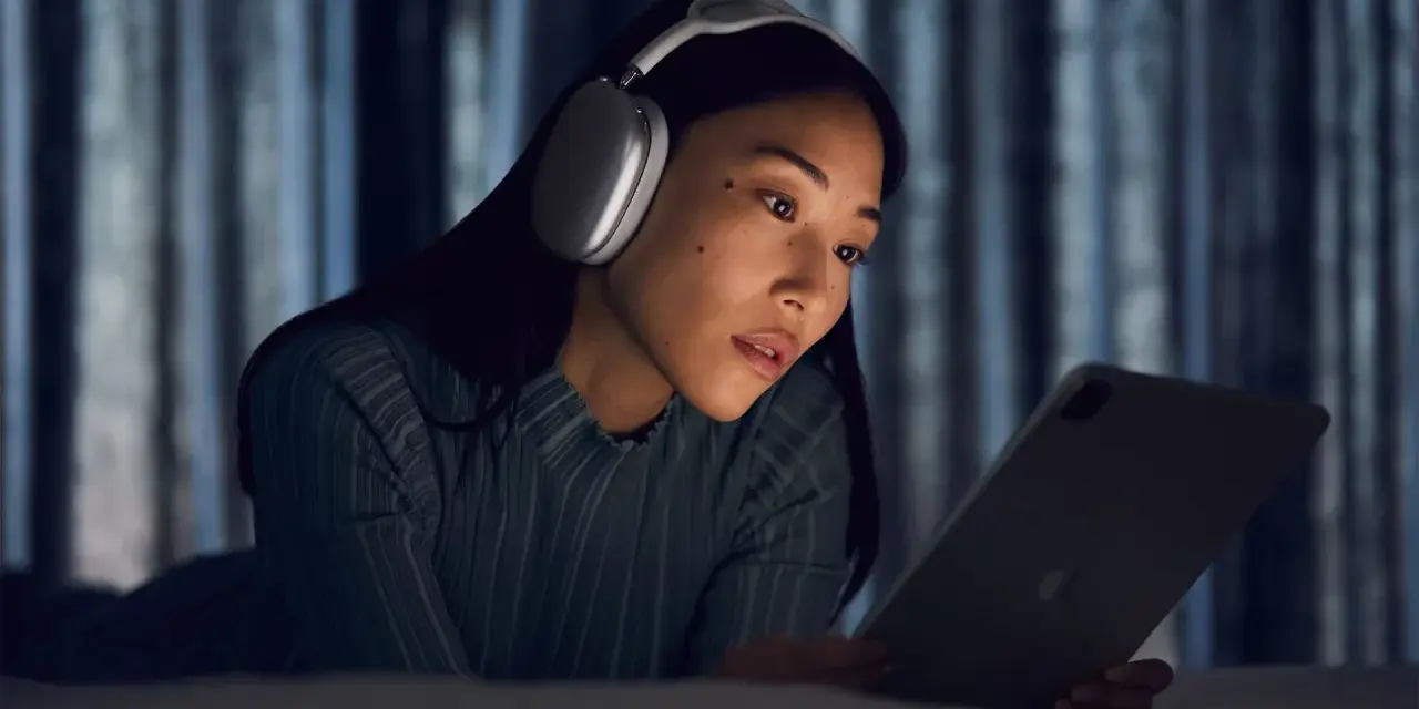 Kobieta w słuchawkach AirPods Max leży na łóżku i patrzy na tablet.