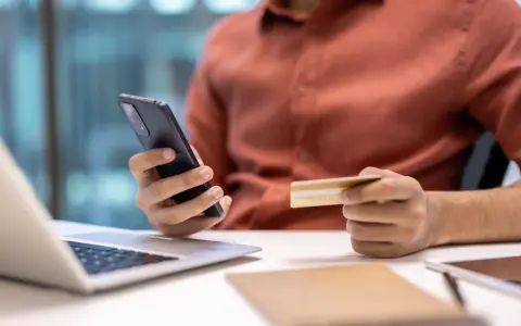 Dłoń trzymająca telefon i kartę kredytową przy laptopie.