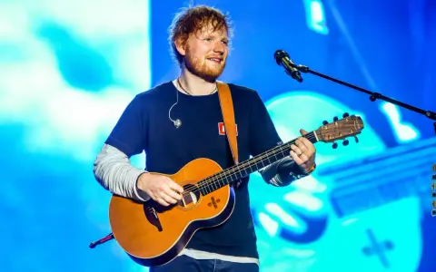 Ed Sheeran gra na gitarze akustycznej na tle niebieskiej sceny.