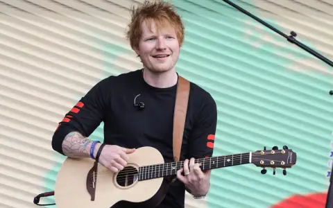 Ed Sheeran gra na gitarze akustycznej na tle pofalowanej, kolorowej kurtyny.
