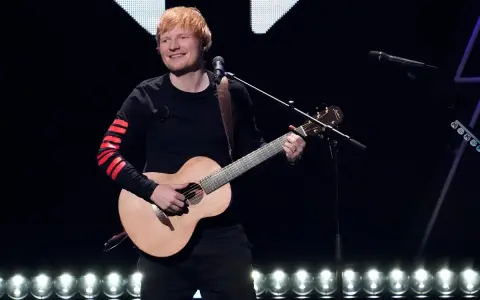 Ed Sheeran gra na gitarze akustycznej na scenie, ubrany w czarną bluzę z czerwonymi pasami na rękawach.