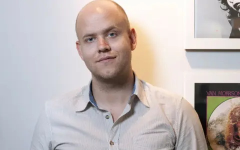 Daniel Ek, założyciel Spotify, w koszuli w paski na tle ściany z plakatami.