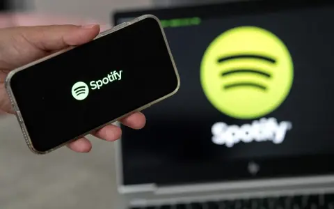 Dłoń trzyma smartfon z logo Spotify. W tle ekran laptopa z tym samym logo.
