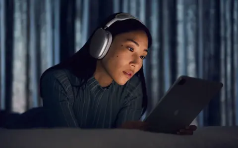 Kobieta w słuchawkach AirPods Max leży na łóżku i patrzy na tablet.
