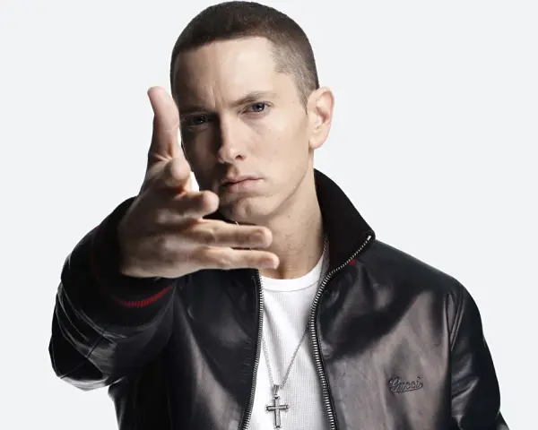 Eminem w czarnej skórzanej kurtce i białej koszulce, z ręką wyciągniętą w stronę widza.