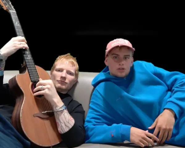 Ed Sheeran z gitarą i młody mężczyzna w niebieskiej bluzie siedzą na kanapie.