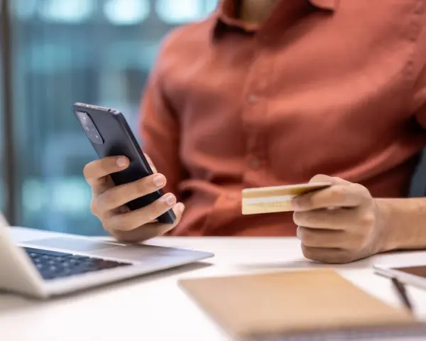 Dłoń trzymająca telefon i kartę kredytową przy laptopie.