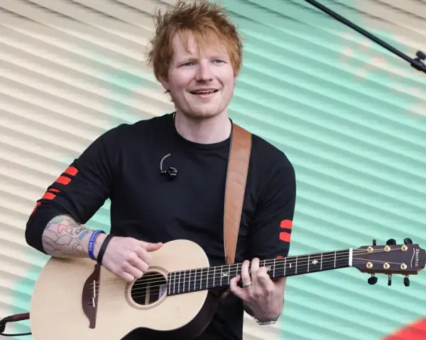 Ed Sheeran gra na gitarze akustycznej na tle pofalowanej, kolorowej kurtyny.