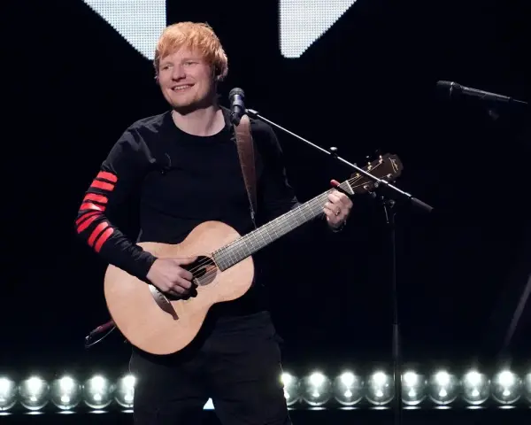 Ed Sheeran gra na gitarze akustycznej na scenie, ubrany w czarną bluzę z czerwonymi pasami na rękawach.