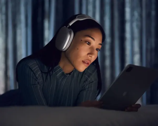 Kobieta w słuchawkach AirPods Max leży na łóżku i patrzy na tablet.