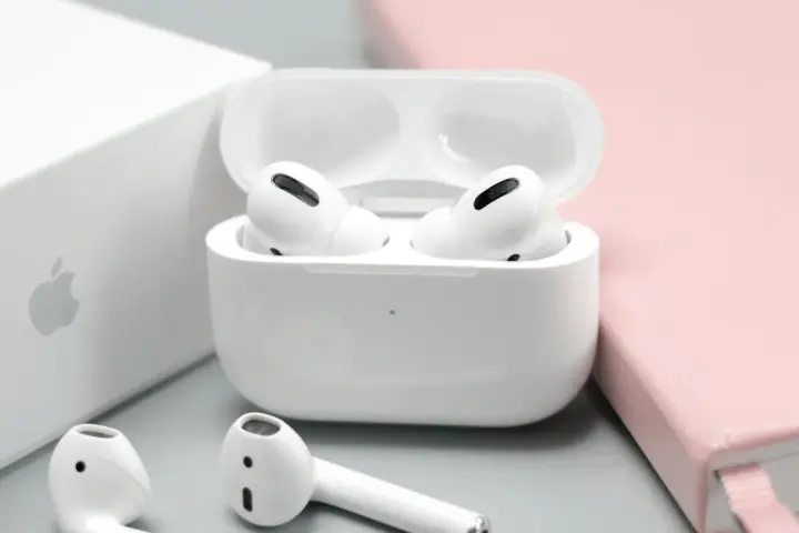 Białe słuchawki AirPods w etui, obok pudełka Apple i różowego notatnika.
