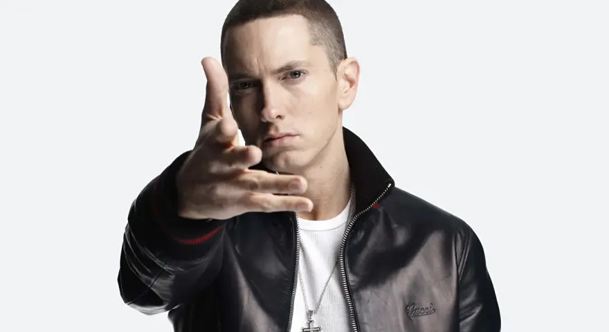 Eminem w czarnej skórzanej kurtce i białej koszulce, z ręką wyciągniętą w stronę widza.