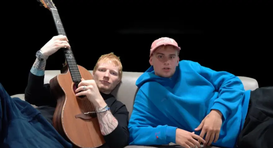 Ed Sheeran z gitarą i młody mężczyzna w niebieskiej bluzie siedzą na kanapie.