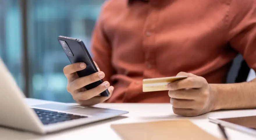 Dłoń trzymająca telefon i kartę kredytową przy laptopie.