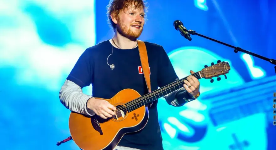 Ed Sheeran gra na gitarze akustycznej na tle niebieskiej sceny.