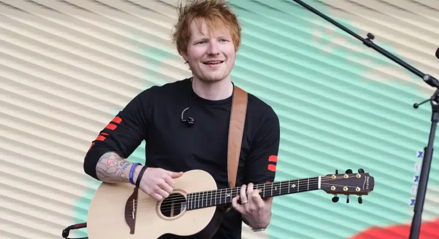 Ed Sheeran gra na gitarze akustycznej na tle pofalowanej, kolorowej kurtyny.
