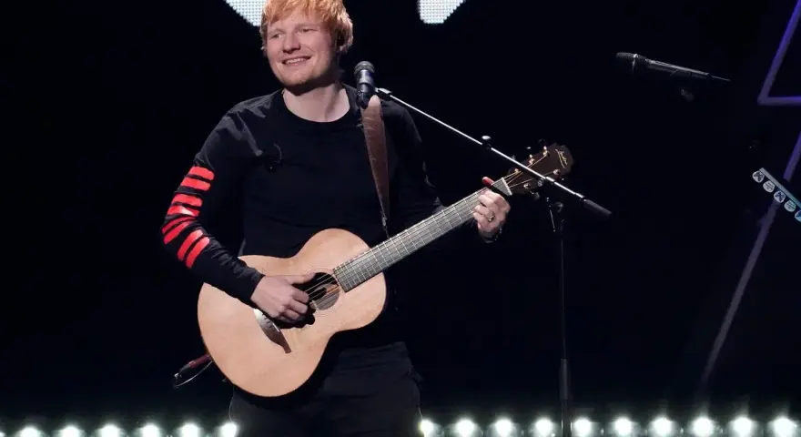 Ed Sheeran gra na gitarze akustycznej na scenie, ubrany w czarną bluzę z czerwonymi pasami na rękawach.
