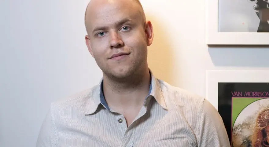 Daniel Ek, założyciel Spotify, w koszuli w paski na tle ściany z plakatami.